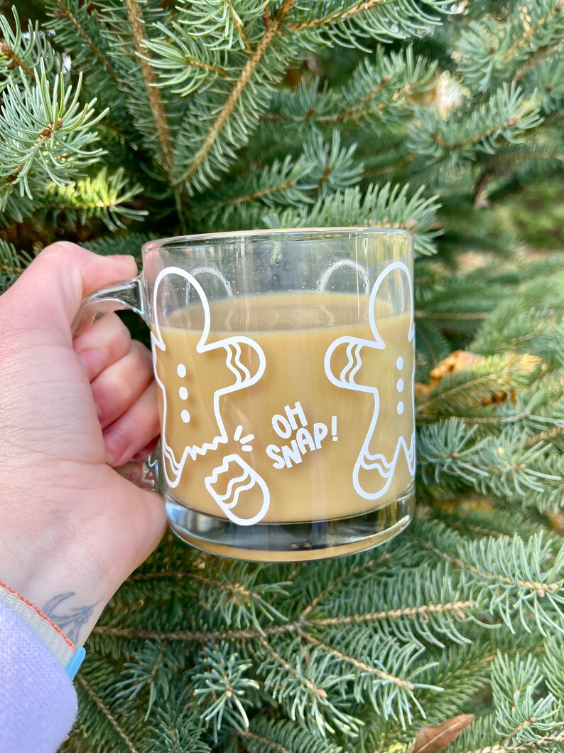 May include: A clear glass mug with a white gingerbread man design. The mug is filled with a brown liquid and has the text "Oh Snap!" on the front.
