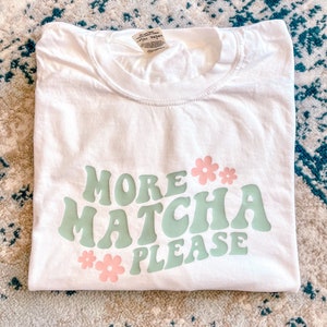 Puede incluir: Camiseta blanca con el texto "More Matcha Please" en letras estilizadas verdes y rosas. El texto est&aacute; rodeado de flores rosas.