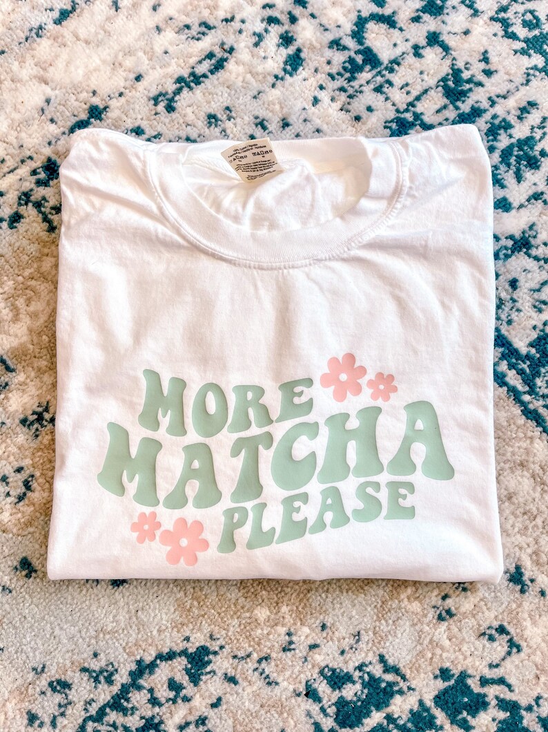 Puede incluir: Camiseta blanca con el texto "More Matcha Please" en letras estilizadas verdes y rosas. El texto est&aacute; rodeado de flores rosas.