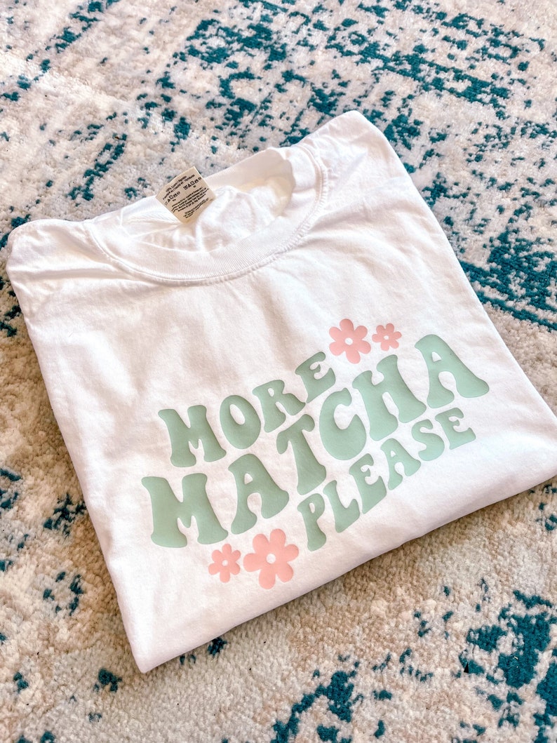 Puede incluir: Camiseta blanca con un gr&aacute;fico verde y rosa que dice "More Matcha Please" con dos flores rosas a cada lado del texto.