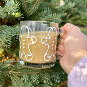 May include: A glass mug with a gingerbread man design, filled with a light brown beverage. The mug is being held by a hand in front of a green evergreen tree.