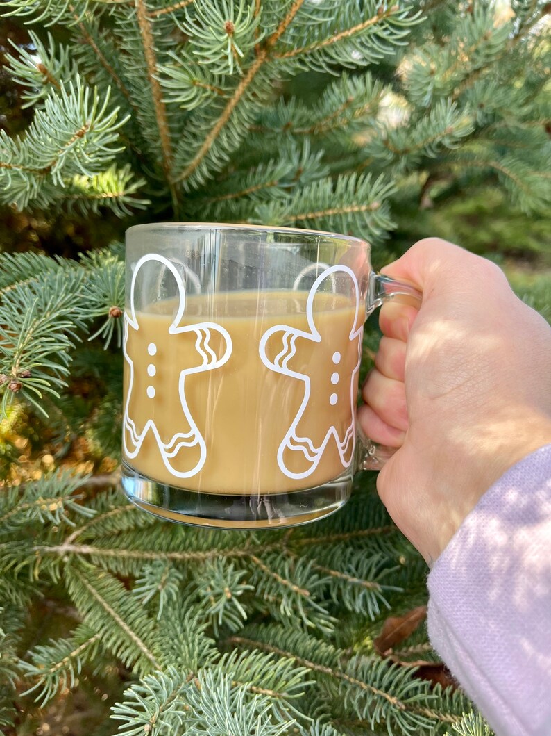 May include: A glass mug with a gingerbread man design, filled with a light brown beverage. The mug is being held by a hand in front of a green evergreen tree.