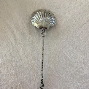 Puede incluir: Una cuchara plateada con un cuenco en forma de concha y un mango largo y ornamentado. El mango presenta un diseño detallado con una pequeña figura en la base. La cuchara es probablemente vintage o antigua, y es adecuada para servir o exhibir.