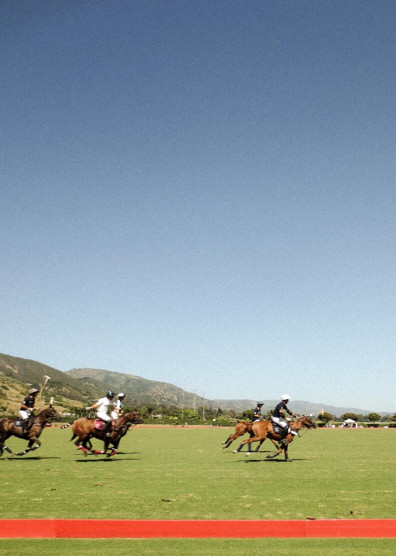 K&ouml;nnte beinhalten: Eine Gruppe von Menschen auf Pferden, die Polo auf einem grasbewachsenen Feld spielen. Die Pferde sind braun und die Reiter tragen wei&szlig;e Hemden und Helme. Der Himmel ist blau und es gibt eine rote Linie auf dem Boden.