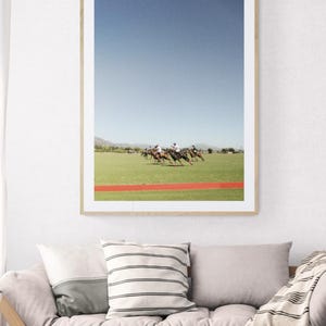 K&ouml;nnte beinhalten: Ein gerahmtes Foto eines Polospiels. Die Spieler reiten auf Pferden und befinden sich mitten im Spiel. Der Himmel ist blau und das Gras ist gr&uuml;n. Es gibt eine rote Linie auf dem Feld.