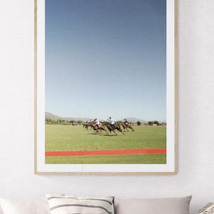 K&ouml;nnte beinhalten: Ein gerahmtes Foto eines Polospiels. Das Bild zeigt mehrere Spieler auf Pferden, die &uuml;ber ein gr&uuml;nes Feld mit einer roten Bahn im Vordergrund reiten. Der Himmel ist strahlend blau.