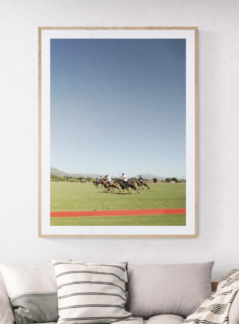 K&ouml;nnte beinhalten: Ein gerahmtes Foto eines Polospiels. Das Bild zeigt mehrere Spieler auf Pferden, die &uuml;ber ein gr&uuml;nes Feld mit einer roten Bahn im Vordergrund reiten. Der Himmel ist strahlend blau.