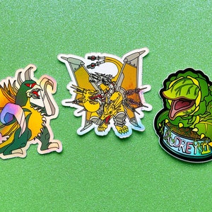 以下が含まれることがあります： 架空の生き物のイラストが描かれた、カラフルなステッカー3枚。1枚目は、虹色のホログラフィックな翼を持つ、緑と黄色のモンスター。2枚目は、鎖と輪を持つ、黄色とオレンジのモンスター。3枚目は、大きな口と「Audrey II」の文字を持つ、緑色の植物モンスター。