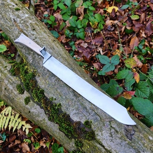 May include: A long, straight-edged knife with a metallic silver blade and a light-colored wooden handle. The knife features a distinct bolster where the blade meets the handle, showcasing a clean, functional design with a sharp, pointed tip.