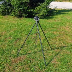 May include: A black metal tripod with a chain and hook, designed for outdoor cooking. The tripod stands on three legs, with a ring at the top for hanging a pot. It is set on a green grassy lawn.