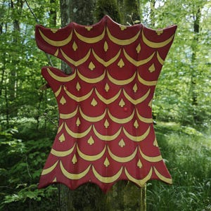 Peut inclure: Un bouclier décoratif rouge et or, monté sur un tronc d'arbre. Le bouclier présente une forme unique et un motif répétitif de formes dorées sur un fond rouge. Le design rappelle des écailles ou un motif stylisé.