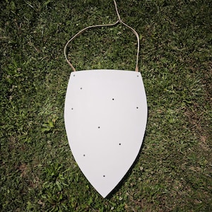Puede incluir: Un escudo de tipo heráldico con una superficie lisa de color gris claro que presenta varios remaches metálicos pequeños y oscuros distribuidos por el frente. Una correa de cuero fina y clara está sujeta al borde superior para colgarlo.