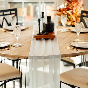 Puede incluir: Un camino de mesa blanco con diseño plisado, colocado sobre una mesa de madera con sillas de metal negro. El camino de mesa está drapeado sobre la mesa y cae hasta el suelo.