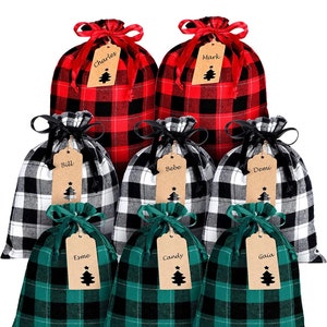 May include: Set of eight Christmas gift bags with plaid fabric in red, black, and white, and green, black, and white. Each bag has a brown paper tag with a name written on it and a small Christmas tree cutout. The names on the tags are Charles, Mark, Bill, Bebe, Demi, Esme, Candy, and Gaia.
