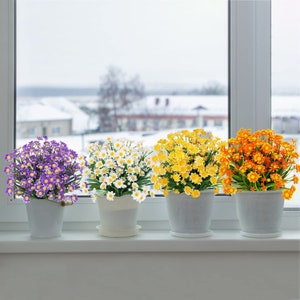 Pode incluir: Quatro arranjos de flores artificiais em vasos brancos. As flores s&atilde;o margaritas brancas, amarelas e roxas.