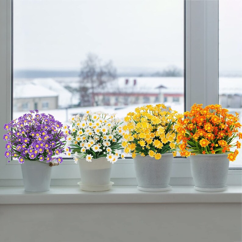 Pode incluir: Quatro arranjos de flores artificiais em vasos brancos. As flores s&atilde;o margaritas brancas, amarelas e roxas.
