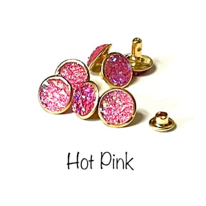 Könnte beinhalten: Nahaufnahme von sechs knallpinken Glitzerknöpfen mit goldenen Einstellungen. Die Knöpfe sind rund und haben eine flache Rückseite. Es gibt auch eine einzelne goldene Einstellung ohne Knopf.