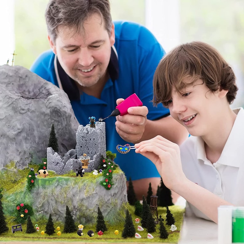May include: A father and son work together on a miniature model landscape. The model features a stone castle, trees, and other details. The father is using a glue gun to attach a small butterfly to the model. The son is holding a small butterfly with tweezers.