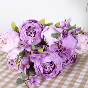 Puede incluir: Un ramo de peon&iacute;as e hortensias artificiales de color p&uacute;rpura con hojas verdes. Las flores est&aacute;n dispuestas en un racimo y est&aacute;n enfocadas. El fondo es una superficie a cuadros de color marr&oacute;n claro y blanco.