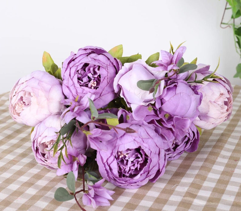 Puede incluir: Un ramo de peon&iacute;as e hortensias artificiales de color p&uacute;rpura con hojas verdes. Las flores est&aacute;n dispuestas en un racimo y est&aacute;n enfocadas. El fondo es una superficie a cuadros de color marr&oacute;n claro y blanco.