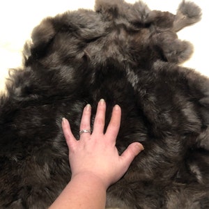 May include: A dark brown sheepskin rug with a soft, fluffy texture. The rug is laid out flat on a white surface.