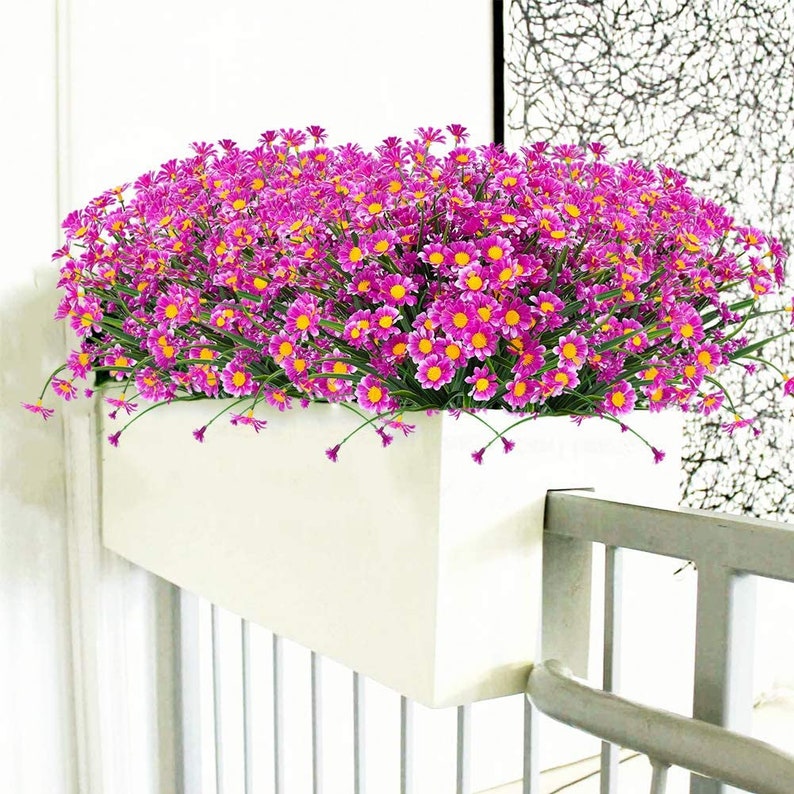 Pode incluir: Um vaso branco com uma grade atr&aacute;s, cheio de um grande buqu&ecirc; de flores artificiais cor-de-rosa.