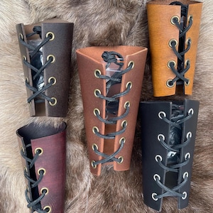 Könnte beinhalten: Fünf Lederarmbänder in verschiedenen Brauntönen, darunter Dunkelbraun, Mittelbraun und Hellbraun. Jedes Armband hat ein Schnürdesign mit schwarzer Kordel und Messingösen. Die Armbänder werden auf einer strukturierten, braunen Oberfläche präsentiert.