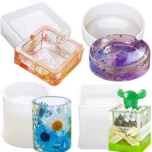 Puede incluir: Cuatro moldes de silicona para hacer manualidades de resina. Los moldes tienen forma de cenicero cuadrado, cenicero redondo, cilindro y caja cuadrada. Los moldes son blancos y se muestran con manualidades de resina terminadas en su interior.
