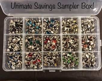 Può includere: Una scatola di contenimento in plastica trasparente con 12 scomparti pieni di strass di vari colori. Gli strass sono fissati a montature in metallo argentato. La scatola è etichettata "Ultimate Savings Sampler Box!"