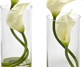 May include: Two clear glass vases with white calla lilies arranged inside. The stems are green and the flowers are white. The vases are filled with water and have a green band around the bottom.