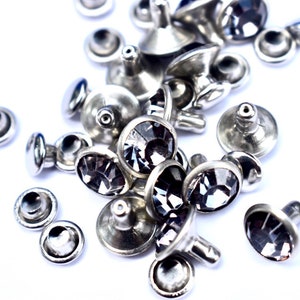 May include: A close-up of a pile of silver-toned metal rivets with black crystal accents. The rivets are scattered on a white surface.