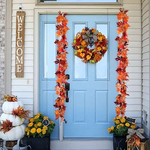 Può includere: Una porta d'ingresso blu con una ghirlanda autunnale e due ghirlande di fogliame autunnale appese su entrambi i lati. La porta ha un cartello di benvenuto bianco a sinistra e una pianta in vaso con fiori gialli a destra.