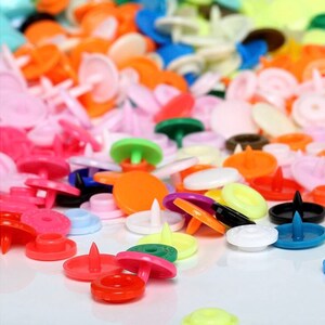 May include: A variety of colorful plastic snap buttons in various shapes and sizes. The buttons are scattered on a white surface.