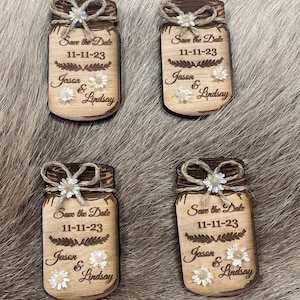 May include: Four wooden save the date magnets shaped like mason jars with twine bows and white flowers. The magnets say "Save the Date" with the date "11-11-23" and the names "Jason & Lindsay".