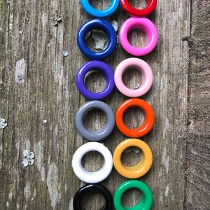 May include: A set of 10 colorful metal eyelets, arranged vertically in a row. The eyelets are in the following colors: blue, red, pink, purple, light pink, gray, orange, white, yellow, and green.