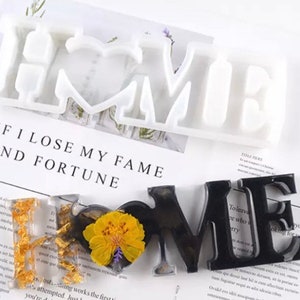 Op de afbeelding: Een witte siliconen mal voor het maken van harsambachten. De mal is gevormd als het woord "HOME" met een hart in de "O". De mal wordt getoond op een wit oppervlak met een stuk papier waarop de tekst "IF I LOSE MY FAME AND FORTUNE" zichtbaar is.