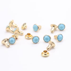 May include: A collection of light blue and gold metal studs with a round, faceted design. The studs are scattered on a white surface.