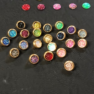 May include: Assortment of small, round, glittery, colorful cabochons with gold settings. The cabochons are arranged in a scattered pattern on a black surface.