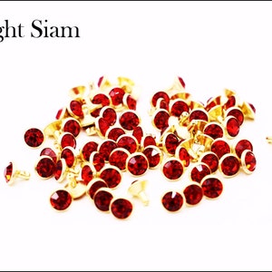 May include: A pile of red rhinestone embellishments with gold settings. The rhinestones are scattered on a white background.