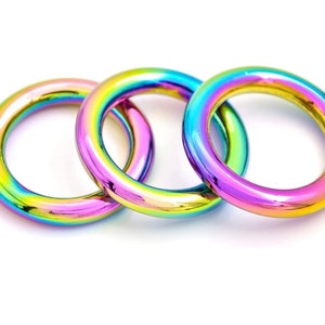 May include: Three iridescent metal rings with a rainbow finish. The rings are circular and have a smooth, reflective surface. The colors shift from yellow to green, blue, and purple. The rings are arranged in a close-up shot against a white background.