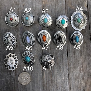 May include: Eleven silver metal conchos with various designs and coloured stones. The conchos are arranged in three rows on a wooden surface. The conchos are labelled A1 through A11.