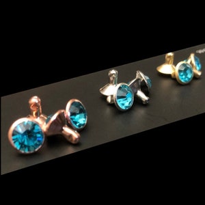 May include: Four small, round, turquoise blue crystal earrings with silver, gold, and rose gold settings. The earrings are arranged in a row on a black surface.