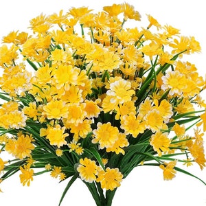 Pode incluir: Um buqu&ecirc; de flores de margarida amarelas artificiais com caules e folhas verdes. As flores est&atilde;o dispostas em um cacho, com algumas das flores voltadas para a c&acirc;mera e outras voltadas para fora.