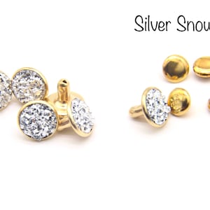 May include: Gold-toned metal studs with a round, silver glitter top. The studs are shown in various positions, including a single stud with a post.