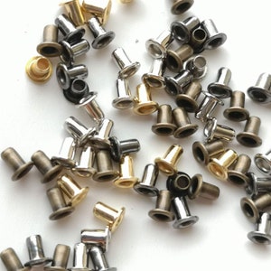 May include: A collection of metal eyelets in gold, silver, and bronze colors. The eyelets are scattered on a white surface.