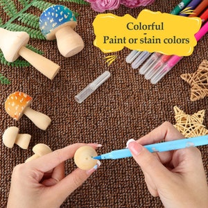 May include: A set of wooden mushroom figurines with colorful paint or stain colors. The mushrooms are in various sizes and shapes. A hand is holding a blue marker and painting one of the mushrooms.