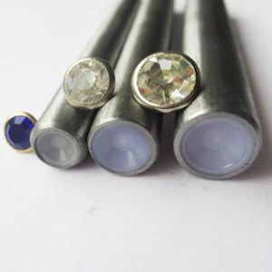 May include: Four metal tools with rubber tips for setting rhinestones. The tools are arranged in a row, with the largest tool on the right and the smallest tool on the left. There are three clear rhinestones and one blue rhinestone.