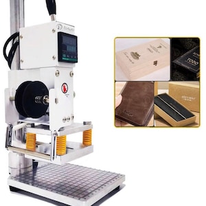 Puede incluir: Una máquina de estampado en caliente eléctrica blanca y plateada con un mango negro y una pantalla digital. La máquina se utiliza para estampar diseños en cuero, madera y otros materiales. La máquina se muestra con una variedad de ejemplos de artículos estampados, incluyendo una caja de madera, una cartera de cuero y una caja negra.