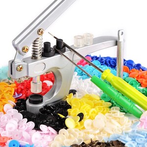 May include: A silver snap press tool with a spring-loaded lever and a black base. The tool is surrounded by a variety of colorful plastic snaps in different sizes and colors, including black, white, yellow, blue, red, and pink.