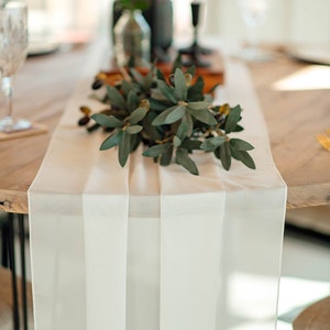 Op de afbeelding: Een witte tafelrunner met een groene bladkrans erop. De runner ligt op een houten tafel.